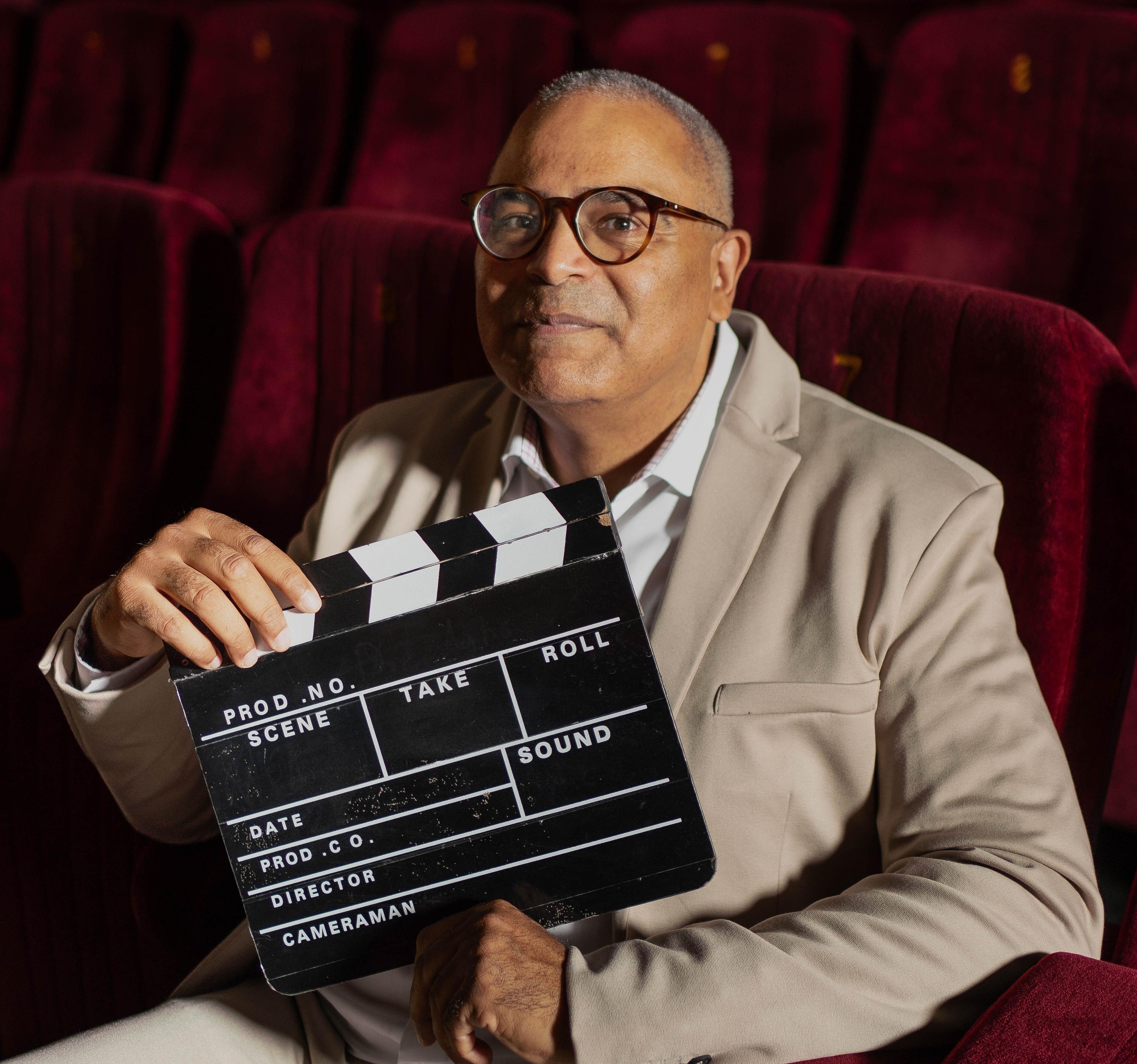 Man in glasses holding a clapperboard while sitting in a movie theater seat.