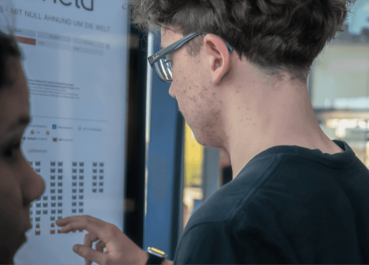 Two young men in glasses examine a digital seating chart screen, one pointing at options.