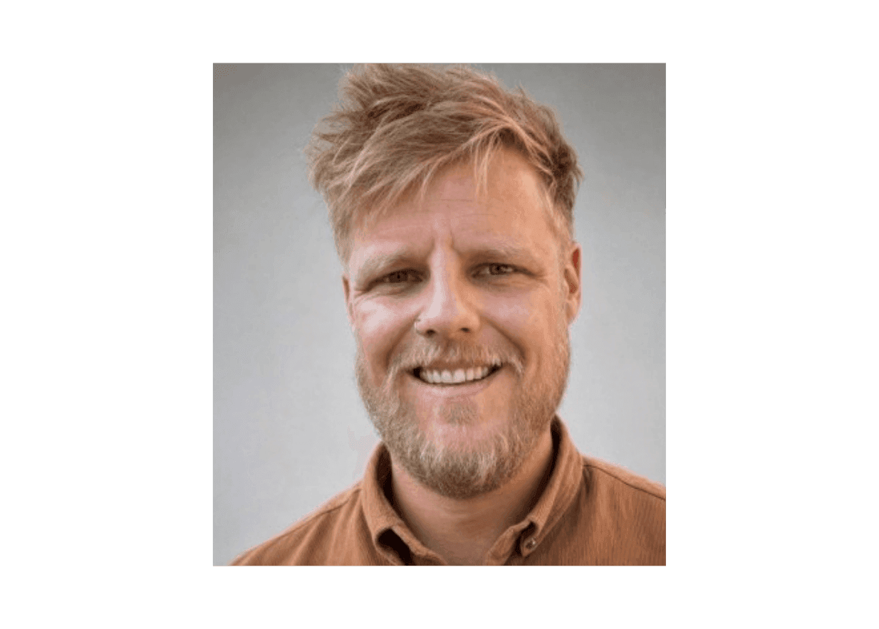 A smiling man with blonde hair and a beard wearing a brown shirt.