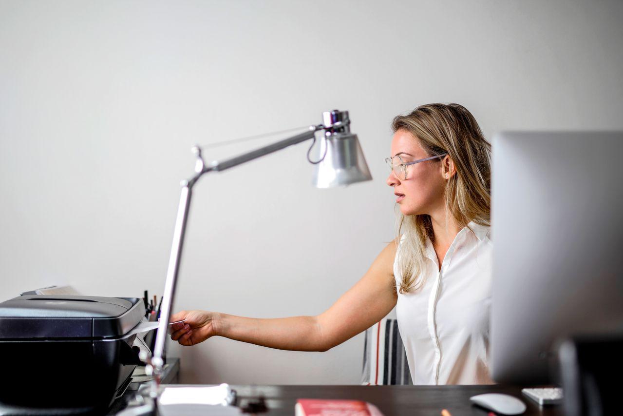 Woman with glasses adjusting a silver desk lamp at her workstation.